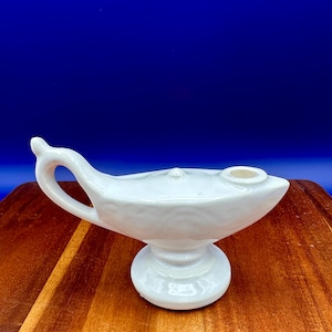 Op de afbeelding: Een witte keramische genielamp met een gebogen handvat en een tuit. De lamp heeft een ronde basis en staat op een houten oppervlak met een blauwe achtergrond.