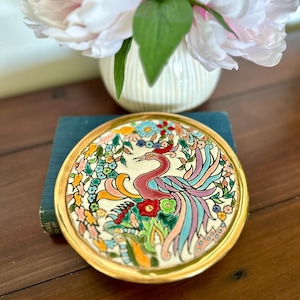 Puede incluir: Un plato decorativo de cerámica con borde dorado, con un diseño de fénix colorido. El ave está representada en tonos de rojo, rosa, azul y verde, rodeada de elementos florales. El plato está sobre un libro azul, con un jarrón de flores rosas al fondo.