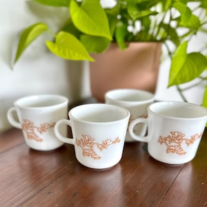 May include: Four white coffee mugs with a floral design in a warm orange hue. Each mug has a handle and a curved shape. The mugs are arranged on a wooden surface, with green plants in the background.