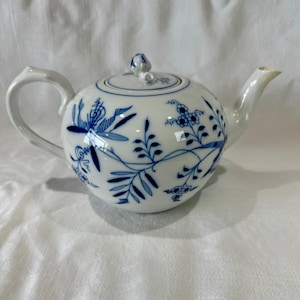 Puede incluir: Tetera de cerámica blanca con cuerpo redondo, asa curva y boquilla. La tetera está decorada con motivos florales y de hojas azules. La tapa tiene un pequeño pomo. La tetera está sobre una superficie blanca.