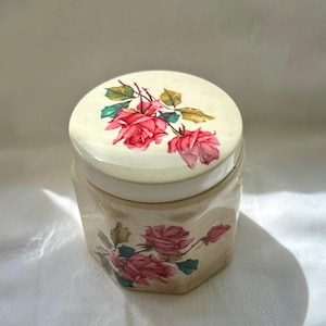 Peut inclure: Un pot hexagonal en céramique beige avec un couvercle, décoré de roses roses et de feuilles vertes. Le couvercle présente un groupe de roses, tandis que le corps du pot présente un motif floral similaire. Le pot est probablement utilisé pour le stockage ou la décoration.