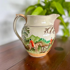 May include: Cream ceramic pitcher with a hunting scene. The pitcher has a handle, spout, and a decorative base. The scene depicts riders on horseback, trees, and a fence. The pitcher is approximately 18 cm tall.