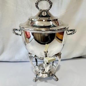 Puede incluir: Una cafetera plateada con tapa y grifo. La cafetera tiene un asa decorativa en la parte superior y dos asas laterales. Se apoya sobre una base decorativa con patas de garra. La cafetera es muy reflectante.