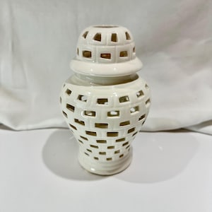 May include: A cream ceramic jar with a lid, featuring a lattice design with rectangular cutouts. The lid also has cutouts. The jar is on a white surface.