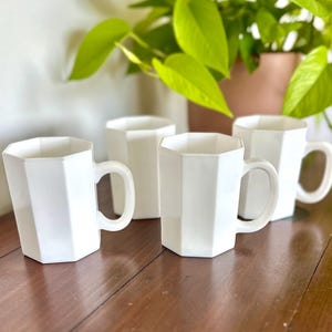 May include: Four white ceramic mugs with geometric, faceted designs. Each mug has a rounded handle. The mugs are arranged on a wooden surface, with a potted plant in the background.