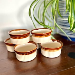 Puede incluir: Un conjunto de seis ramequines vintage. Cada ramequín tiene una base color crema con estrías verticales y un borde marrón. Los ramequines están apilados sobre una superficie de madera oscura, con una planta en el fondo.