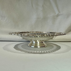 May include: A silver serving dish with a pedestal base. The dish has a wide, shallow bowl with a decorative, scalloped edge. The base is round and fluted. The dish is likely made of metal and reflects light.