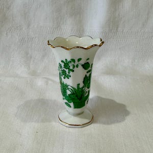 May include: A small, white porcelain vase with a scalloped rim and a gold trim. The vase features a green floral design and a pedestal base, also trimmed in gold. The vase is approximately 6 inches tall.