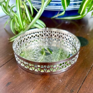 May include: A silver-coloured, round coaster with an openwork geometric design around the edge. The coaster has a reflective surface with a floral pattern in the centre. The coaster is likely made of metal and is designed to protect surfaces from drink rings.