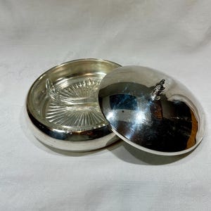 Może przedstawiać: Srebrna patera do serwowania z pokrywką. Naczynie ma okrągłą podstawę z wkładką szklaną i pokrywkę z ozdobnym uchwytem. Pokrywka jest częściowo otwarta, odsłaniając wnętrze naczynia. Naczynie stoi na białej powierzchni.