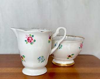 Può includere: Set di lattiera e zuccheriera in ceramica bianca. La lattiera ha un manico e un bordo smerlato. Entrambi i pezzi sono decorati con motivi floreali colorati, tra cui rose rosa e nontiscordardimé blu. La zuccheriera ha un bordo dorato.