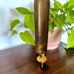 以下が含まれることがあります： 背の高い円筒形のデザインのヴィンテージ真鍮キャンドルホルダー。キャンドルホルダーは、小さな円形のベースと、キャンドル用のカップ型のホルダーを備えています。真鍮は暖かく金色の色合いで、ホルダーは濃い木製の表面に置かれています。