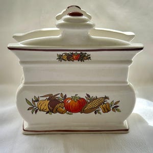 May include: A white ceramic spice jar with a lid, decorated with a band of colourful vegetables including a tomato, corn, and onion. The jar has a rectangular base and a handle on the lid.