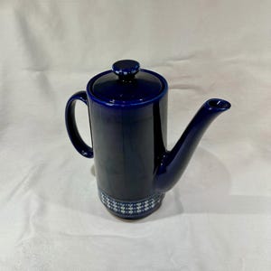 May include: A navy blue ceramic coffee pot with a lid and a spout. The pot has a handle and a decorative band of white and blue geometric patterns near the base. The pot is on a white surface.