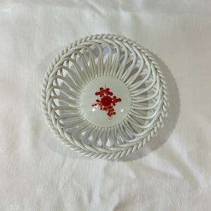 May include: A white, round, woven basket with a decorative floral design in the center. The basket has a braided edge and a central white ceramic disc with a red floral pattern. The basket is likely used for decorative purposes.