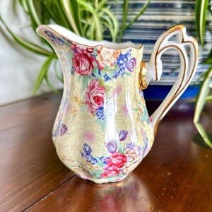 Könnte beinhalten: Ein Milchkännchen aus Porzellan mit hellgelbem Hintergrund, einer feinen Rissstruktur und einem bunten Blumenmuster aus roten, rosa und violetten Blüten. Das Kännchen hat einen gewellten Rand und einen verzierten Henkel mit Goldrand.