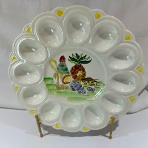 Op de afbeelding: Een witte keramische oester serveerschaal met 12 individuele vakken. Het midden heeft een geschilderd stilleven van een wijnfles, ananas, kaas en druiven. Gele accenten en een geschulpte rand maken het ontwerp compleet. De schaal wordt op een goudkleurige standaard gepresenteerd.