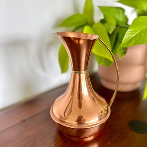 Könnte beinhalten: Eine kupferfarbene Kanne mit ausgestelltem Oberteil und gebogenem Griff. Die Kanne hat einen bauchigen Boden und einen schmalen Hals, mit einem goldfarbenen Band am Hals. Die Kanne steht auf einer dunklen Holzoberfläche.
