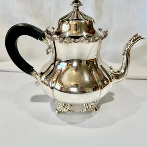 Puede incluir: Una tetera plateada con un asa negra y detalles ornamentados. La tetera tiene una tapa con un adorno decorativo y un pico curvo. La base de la tetera tiene un diseño decorativo con patas. La tetera es muy reflectante.