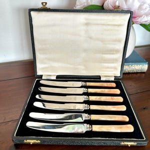 Könnte beinhalten: Ein Set aus sechs antiken Messern in einer schwarzen Box. Die Messer haben silberne Klingen und hellbraune, perlmuttfarbene Griffe. Die Box ist mit cremefarbenem Stoff ausgekleidet.