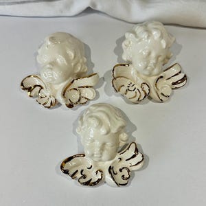 Könnte beinhalten: Drei Wandbehänge aus Keramik mit Cherubim. Jeder Cherub hat ein cremefarbenes Gesicht und Haar mit goldfarbenen Akzenten auf den Flügeln. Die Cherubim sind auf einer weißen Oberfläche angeordnet.