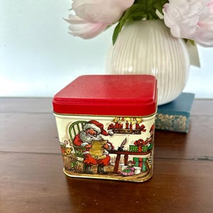 Può includere: Una scatola di Natale vintage con coperchio rosso e corpo color crema. La scatola presenta un'illustrazione colorata di Babbo Natale che legge una lista, circondato da regali e candele. La scatola è di forma quadrata e poggia su una superficie di legno.