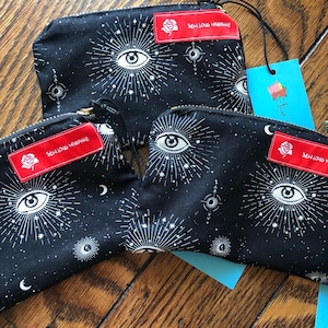 Pode incluir: Bolsas pretas com zíper com um padrão de olho celestial e estrelas. Cada bolsa tem uma etiqueta vermelha com o texto "Sew Long Marianne" e um gráfico de rosa. As bolsas estão em uma superfície de madeira.
