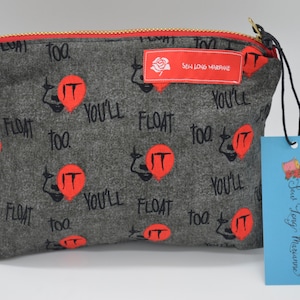 Puede incluir: Bolsa gris con cremallera, interior rojo y cremallera dorada. La bolsa tiene un estampado de globos rojos y las palabras "IT" y "YOU'LL FLOAT TOO" en negro. Una etiqueta azul con "Sew Long Marianne?" cuelga de la cremallera.
