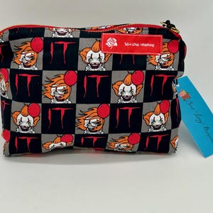 Peut inclure: Une trousse de maquillage à carreaux noirs et gris avec un motif répété d'un clown avec des ballons rouges et le mot "IT" imprimé dessus. La trousse a une fermeture éclair et une petite étiquette qui indique "Sew Long Marianne".