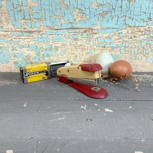 May include: A vintage stapler with a cream-colored body and red accents, along with a box of Arrow staples. The stapler is positioned next to a white egg and a brown onion, with a rustic wooden background.
