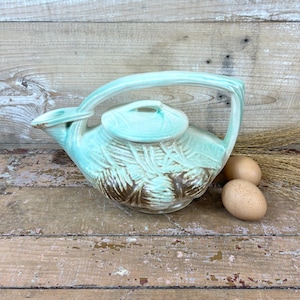 May include: A light mint green ceramic teapot featuring a raised relief pattern of pinecones and needles. The teapot has a curved handle that extends from the spout to the back, a matching lid, and a glossy finish with brown accents on the pinecones.