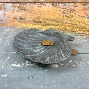 May include: A dark, weathered metal dish shaped like a scallop shell with a textured, ridged surface. The finish appears aged with hints of dark gray and bronze tones. Two copper-colored pennies rest inside the shell, and one sits beside it.