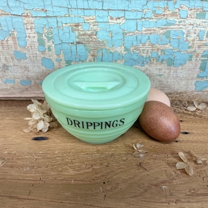 May include: A vintage, light green glass bowl with a lid, labeled "DRIPPINGS" in black lettering. The bowl has a ribbed design and is accompanied by two brown eggs. The background features weathered wood and dried flowers.