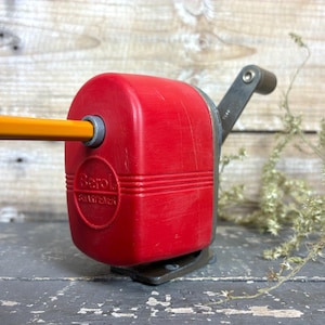 May include: A red Berol manual pencil sharpener with a grey crank handle. A yellow pencil is inserted into the front opening. The sharpener features a rectangular body with horizontal ridges and the embossed text Berol SHARPENER on the front.