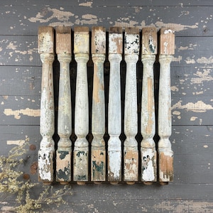 May include: A collection of nine weathered wooden architectural columns. The columns are painted in shades of white, gray, and brown, with visible signs of wear and peeling paint. The columns are arranged on a distressed gray wooden surface.