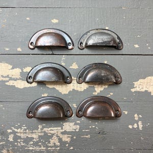May include: Six antique, half-moon drawer pulls with a distressed, aged appearance. The metal pulls have a dark, tarnished finish with visible wear and tear, suggesting they are vintage or reclaimed hardware. They are arranged on a weathered, gray wooden surface.