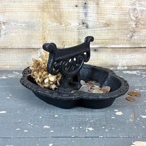 May include: Black cast iron decorative dish with a well-like design, holding coins and dried flowers. The dish has a scalloped edge and a decorative handle. The background is a weathered wooden surface.