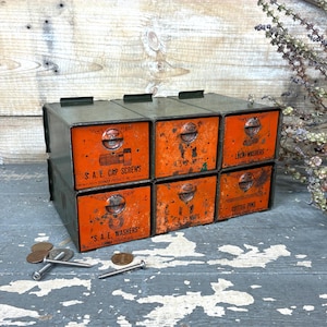 May include: A set of six vintage metal storage drawers. The drawers are orange with black text that reads "S.A.E. CAP SCREWS", "U.S. NUTS", "LOCK WASHERS", "S.A.E. WASHERS", "E. NUTS", and "COTTER PINS".