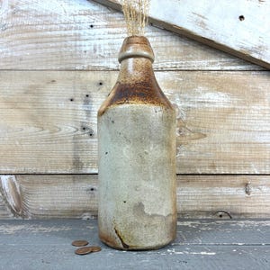 May include: A vintage stoneware bottle with a brown and tan glaze. The bottle has a cylindrical shape with a narrow neck and is filled with dried grasses. Three pennies are at the base of the bottle.