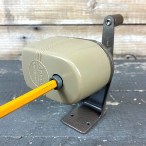 May include: A vintage Apsco pencil sharpener with a beige plastic casing and a dark brown metal base. The sharpener features a manual hand crank and a circular opening for inserting a yellow pencil. The brand name Apsco is embossed on the front.