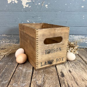 May include: A vintage wooden Edison Lamp Box with a rectangular shape and a handle cutout. The box is made of light brown wood and has the words "EDISON LAMP BOX" printed on the front. Two eggs and dried flowers are next to the box.