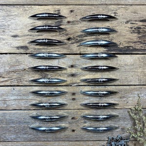 May include: A collection of 16 silver-toned cabinet pulls arranged on a weathered wooden surface. Each pull has a curved, elongated design with a decorative emblem in the center. The pulls are evenly spaced, creating a symmetrical pattern.