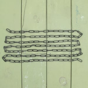 May include: Several lengths of dark metal chain are arranged horizontally against a light green wooden background. The chains are made of interlocking oval links. The wood has vertical planks with visible grain and some imperfections.
