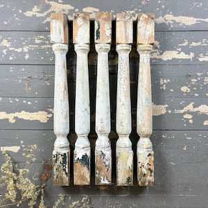 May include: Five antique wooden balusters with a distressed, weathered appearance. The balusters are painted white with visible cracking and peeling paint, revealing the natural wood underneath. They are arranged vertically against a gray wooden background.