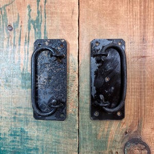 May include: Two rectangular black metal door handles with a weathered, textured finish. Each handle features a central U-shaped pull attached to a flat mounting plate with four circular screw holes at the corners, showing signs of wear and oxidation.