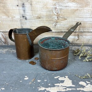 May include: Two antique copper kitchenware items: a pitcher with a spout and a small pot with a handle. The pitcher has a handle and a spout. The pot has a handle and a green interior. Both items show signs of age and use.