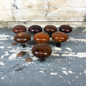 May include: A collection of eight vintage wooden cabinet knobs in various shades of brown, from dark mahogany to light amber. The knobs have a rounded, mushroom-like shape and are arranged on a weathered, gray-blue wooden surface.