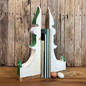 May include: A pair of white and green painted wooden bookends with a curved, pointed silhouette. The bookends are positioned on either side of three upright books with gold and black spines, accompanied by one brown and one light blue egg.