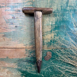 May include: A vintage wooden garden tool with a T-shaped handle and a metal tip. The tool is brown and shows signs of wear. The background is a weathered wooden surface with shades of brown and teal.