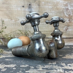 May include: Two antique silver-toned faucet handles with cross-shaped knobs. The faucets are connected by a pipe and sit on a weathered, gray surface. A blue and brown egg are in the background.